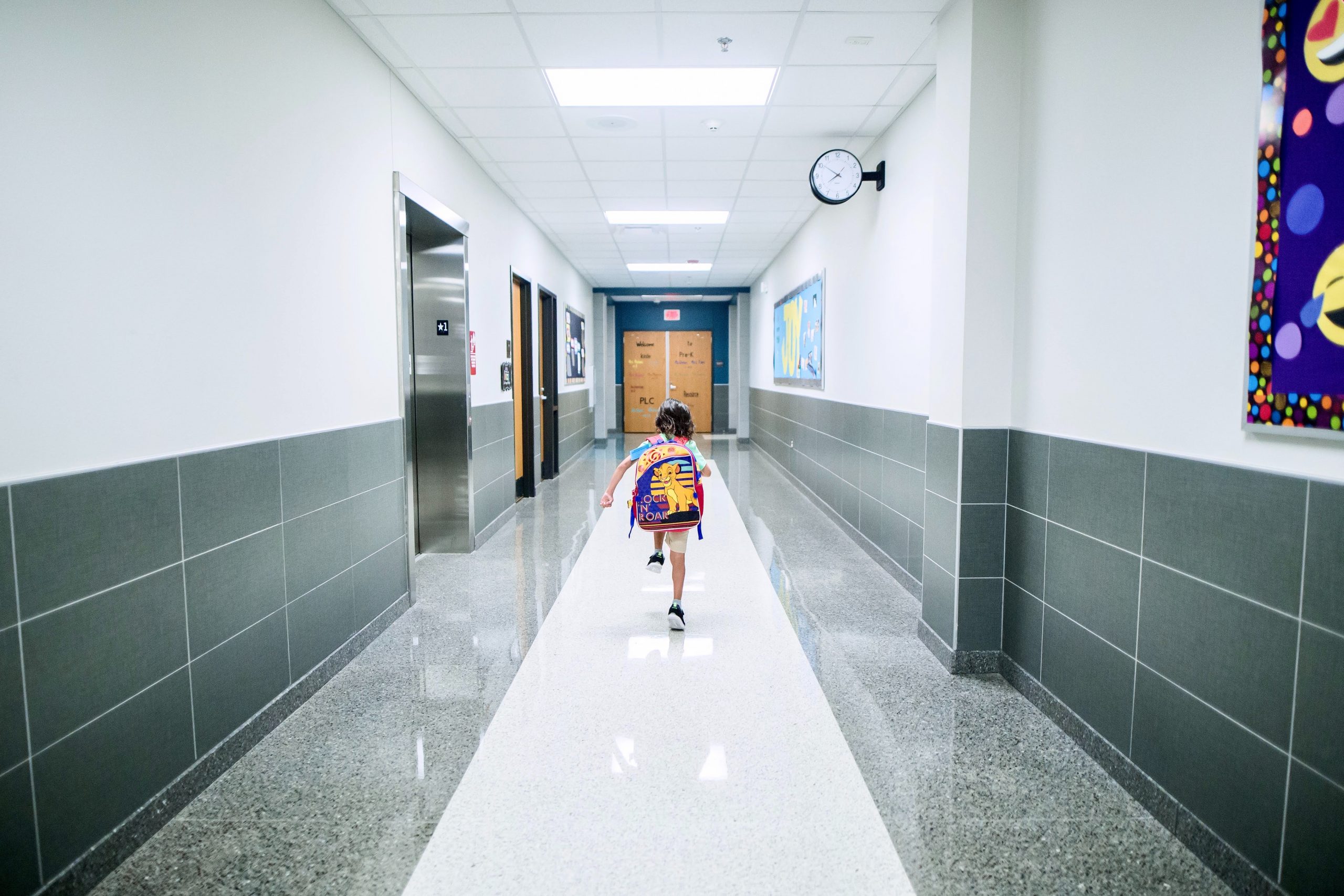 A kid walking down a big school hallway wearing a large backpack.